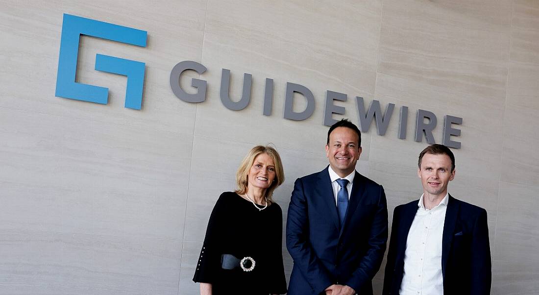 Three people stand in front of a wall that has the Guidewire logo on it.