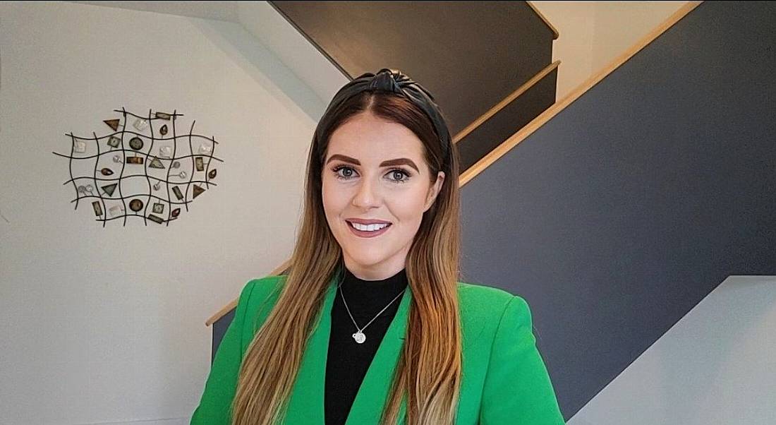 A young woman wearing a green blazer stands in a bright room. She is smiling at the camera.