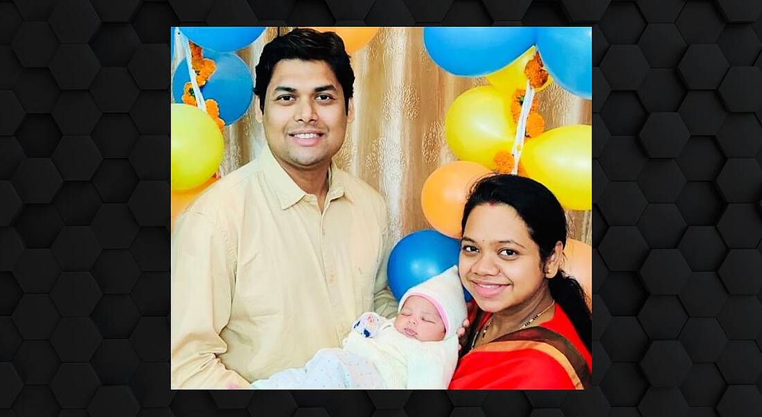 A man and a woman smile at the camera while holding a newborn baby. There are balloons in the background.