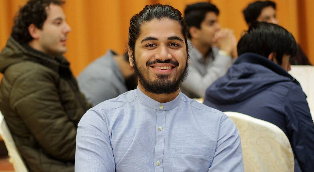 A man wearing a blue shirt smiling at the camera. Behind him are several other people sitting around.