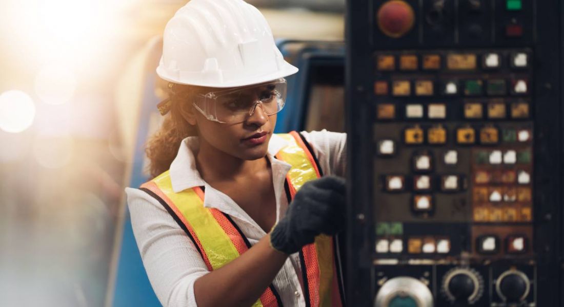 A woman engineer wearing a high-vis jacket and hardhat while working on a factory machine.