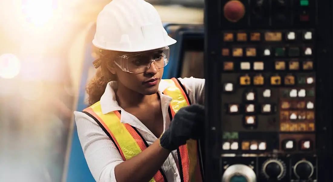 A woman engineer wearing a high-vis jacket and hardhat while working on a factory machine.