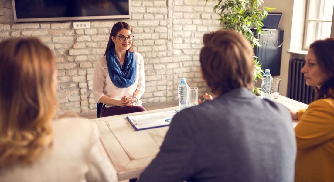 A woman sits across from three other people in a job interview. The office is bright and there’s a plant in the corner.