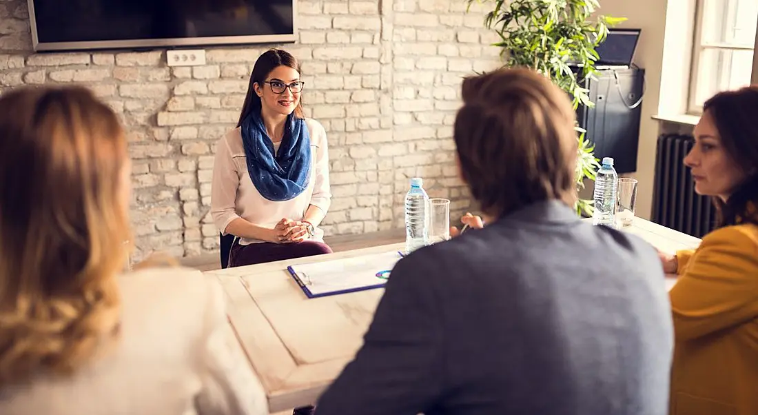 A woman sits across from three other people in a job interview. The office is bright and there’s a plant in the corner.