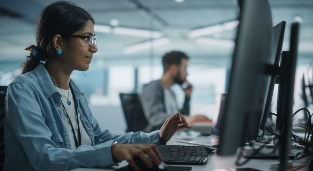 A young woman working at a computer in an office, with a man in the background also working at a computer. They are software engineers.