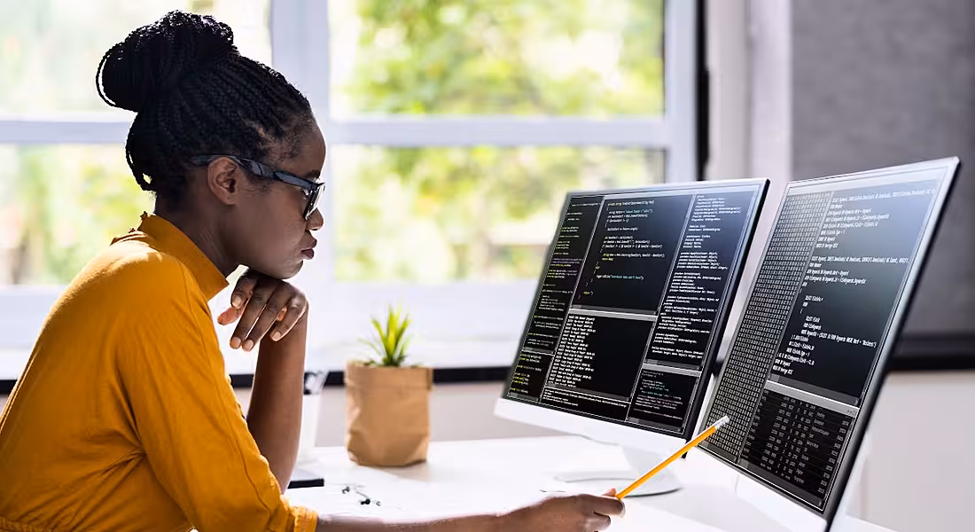 A young woman sits at a desk in a brightly lit room. She’s looking at two large screens with code on them.