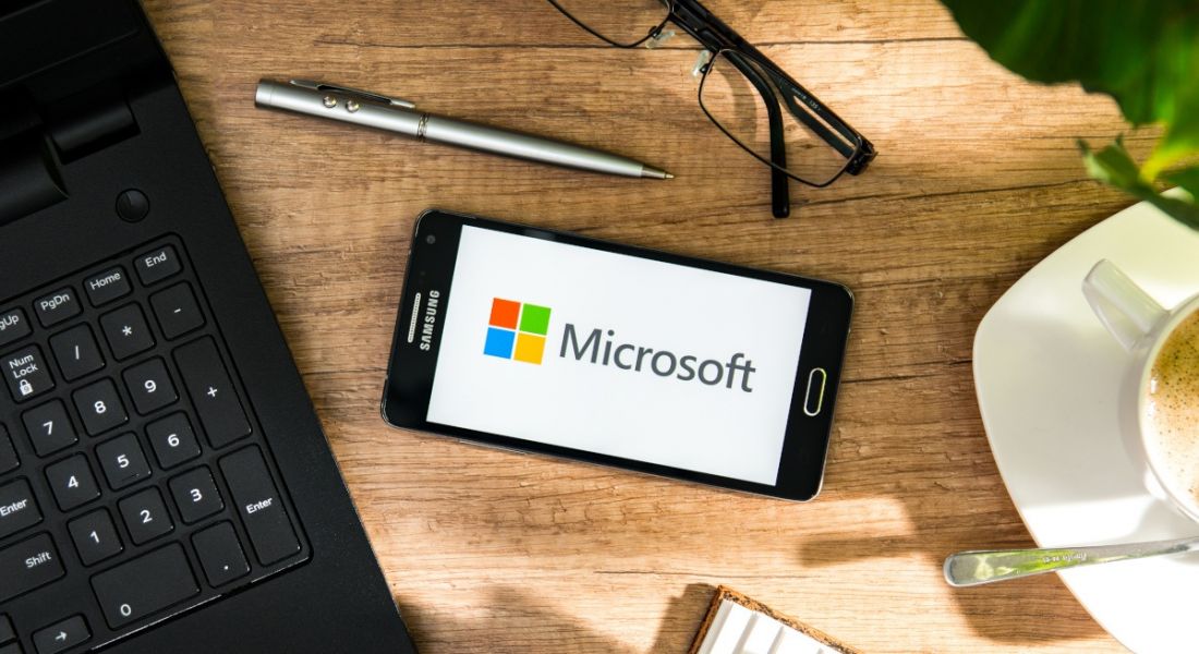 Microsoft's logo displayed on a phone lying on a hybrid worker's desk with a coffee cup, pen, glasses and a laptop.