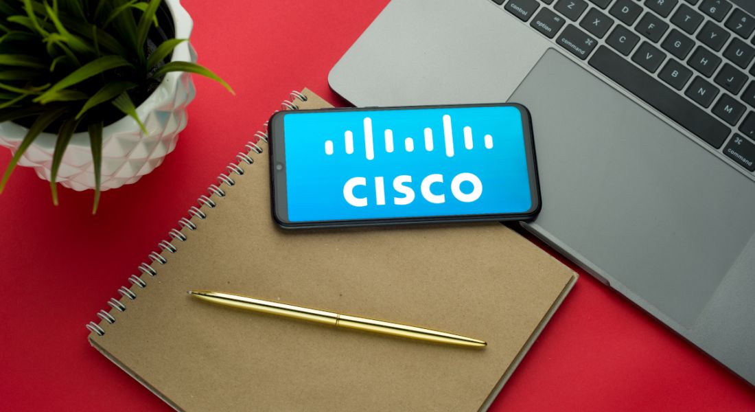 A phone with the Cisco logo on it lying on a notebook with a laptop, pen and pot plant.
