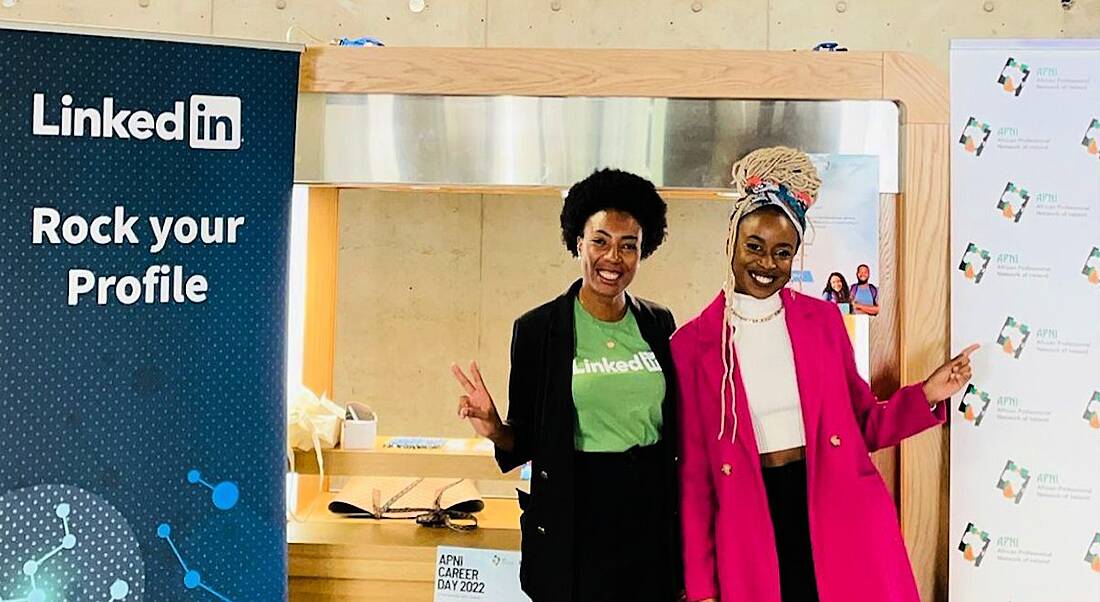 Two women from the African Professional Network of Ireland stand together smiling at the camera. There is a LinkedIn poster beside them.