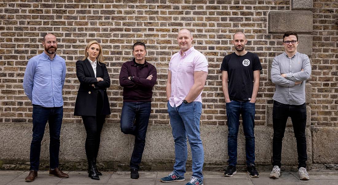 One woman and five men standing next to each other in front of a brown brick wall. They are part of the Gigable team.