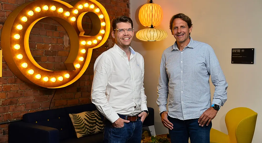 Two men standing in a room with light fixtures and a red brick wall behind them.