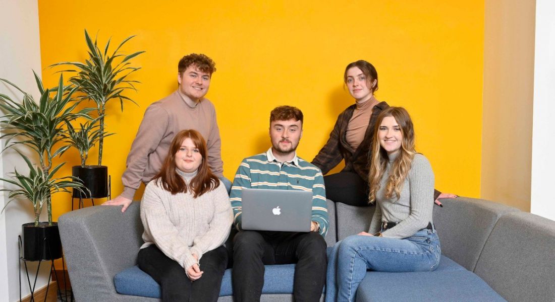 Five people from Liberty IT gathered around a sofa in a room with a yellow feature wall and a pot plant.