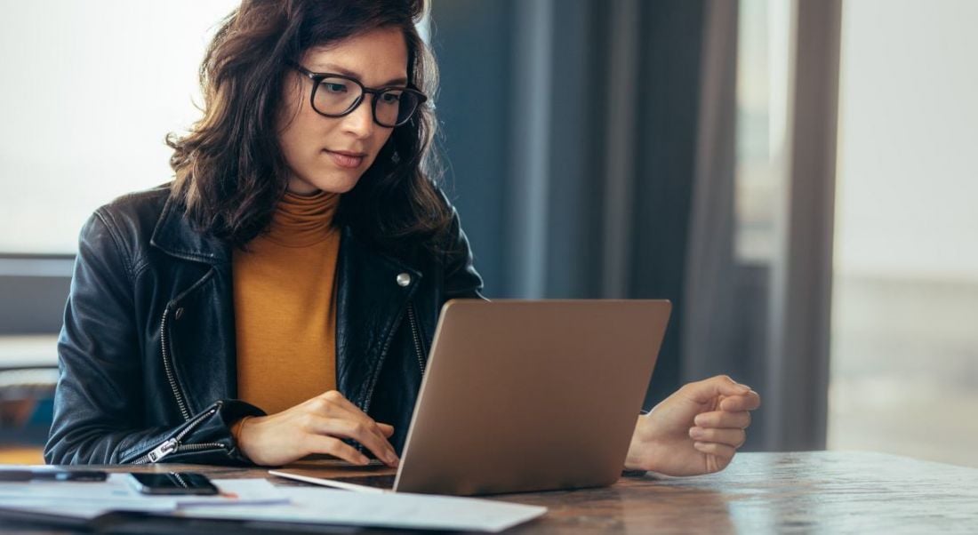 A young woman wearing glasses sits at a desk working on a laptop.