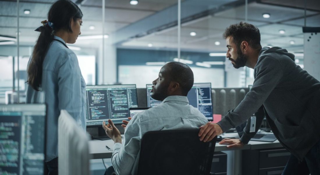 A man and a woman stand either side of another man sitting in a chair at group of screens showing code, symbolising cybersecurity roles.