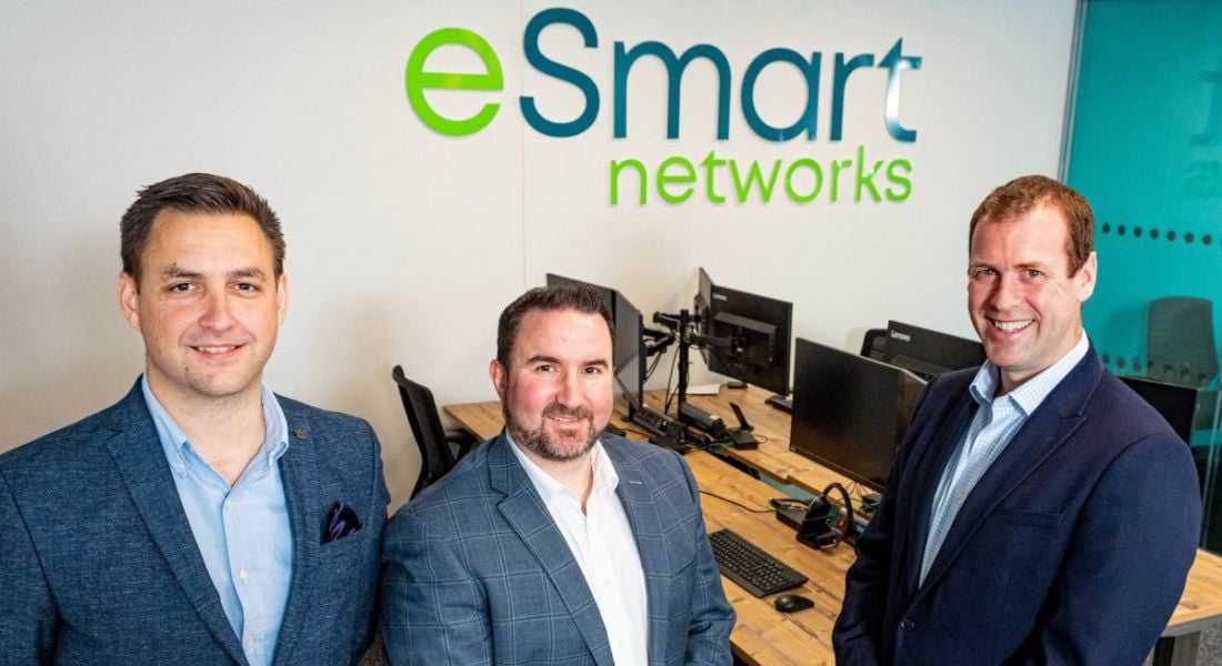 Three men from eSmart Networks and Invest NI standing in an office with eSmart's logo on a board behind them.