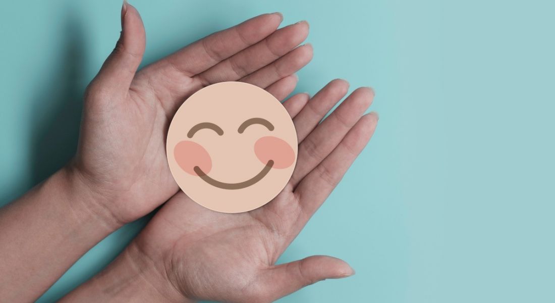 Hands holding a flat smiley face against a blue background, symbolising workplace wellbeing.