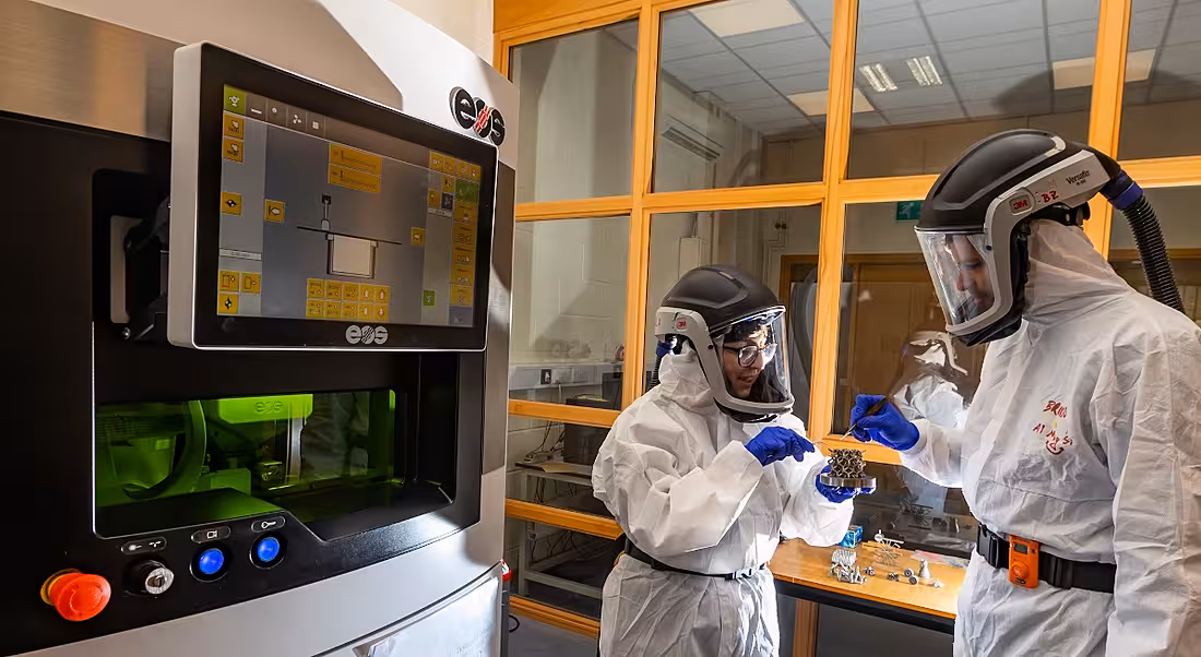 Two people dressed in protective gear as they work with a 3D printer.