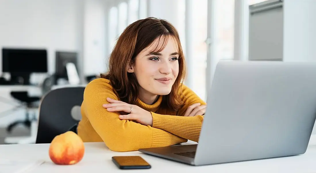 A woman at work is sitting, smiling at her laptop.