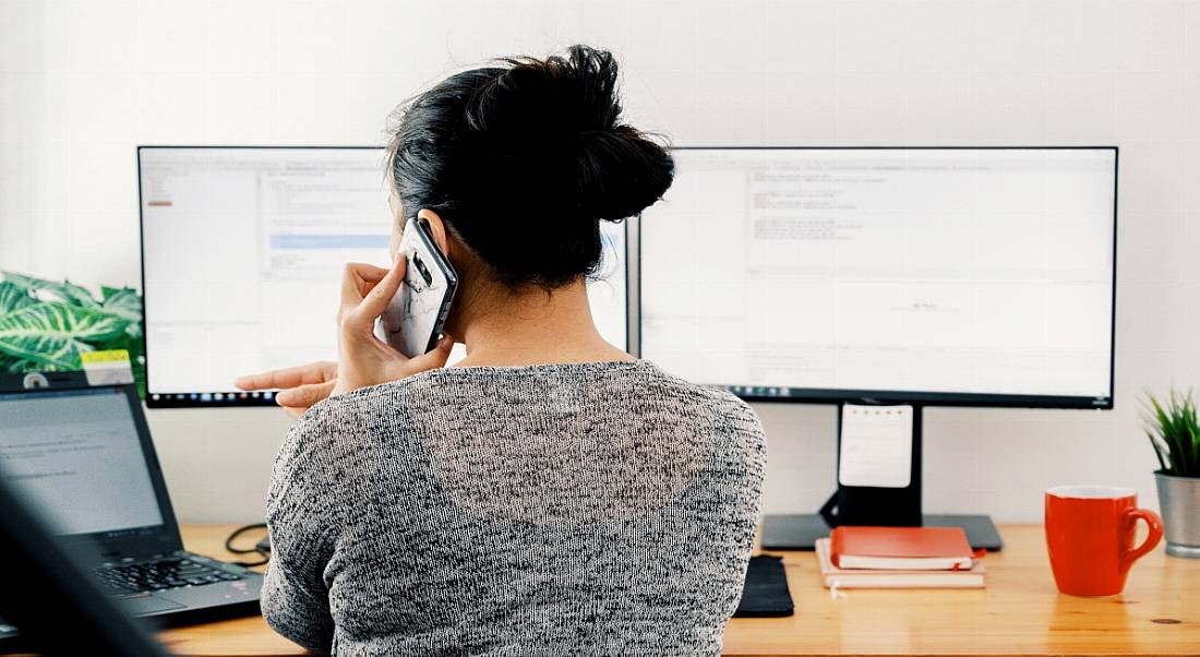 Woman working at home with two computer monitors and a cup beside her.