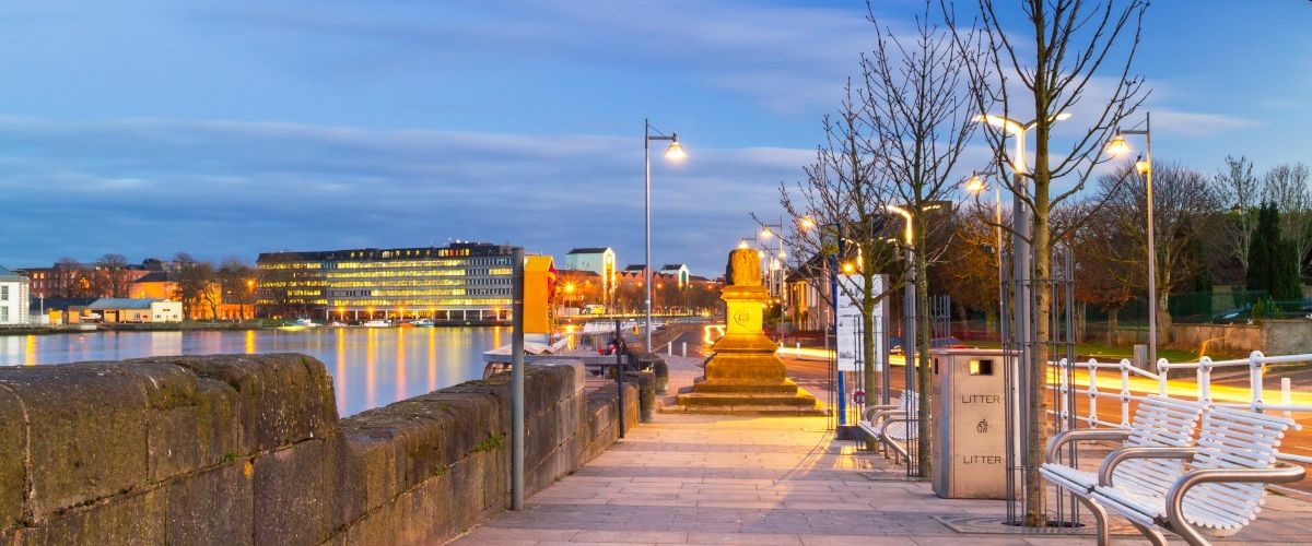 The River Shannon walkway lit up by streetlights in the evening with Limerick city visible across the river.