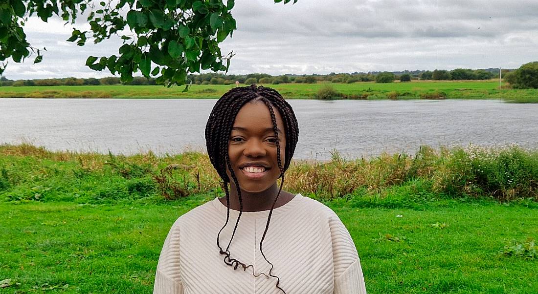 A young woman wearing a cream jumper standing outdoors against a scenic lake. She is smiling at the camera.