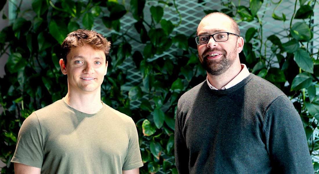 Two men standing against a wall with foliage. They are smiling at the camera.