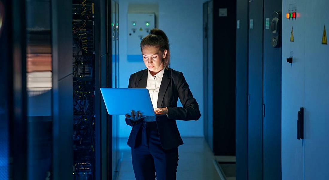 A woman standing in the middle of a network server room while holding a laptop she’s working on.