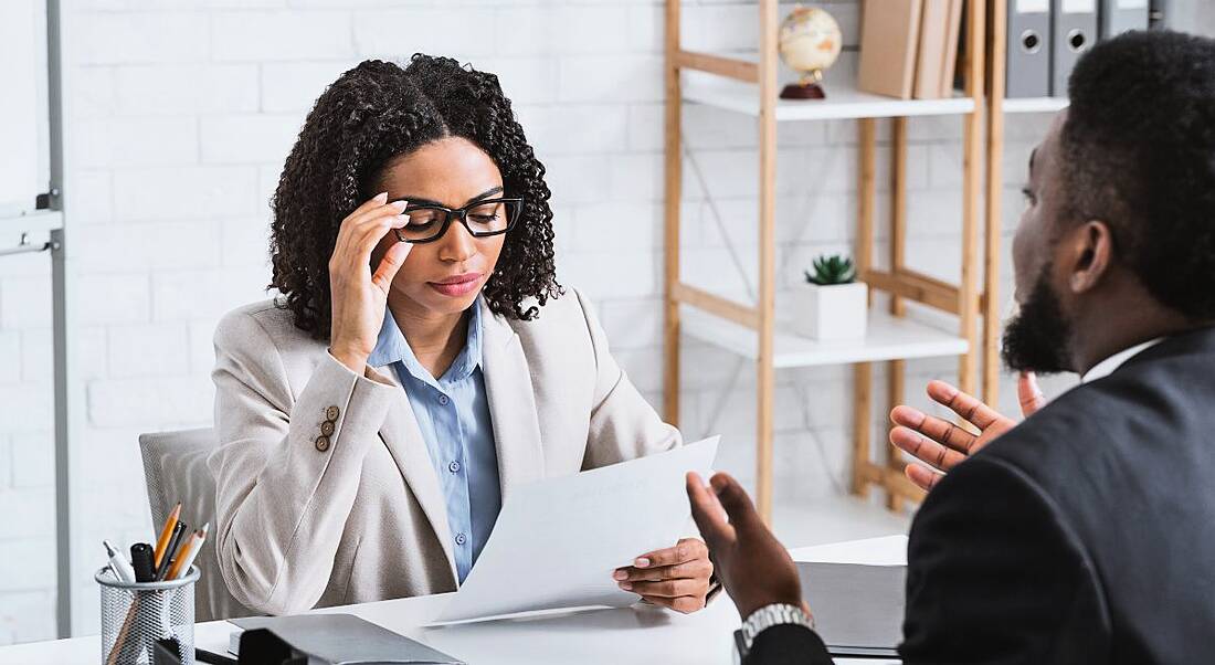 A man and a woman sit on opposite sides of a desk. The woman is inspecting his CV in a job interview.