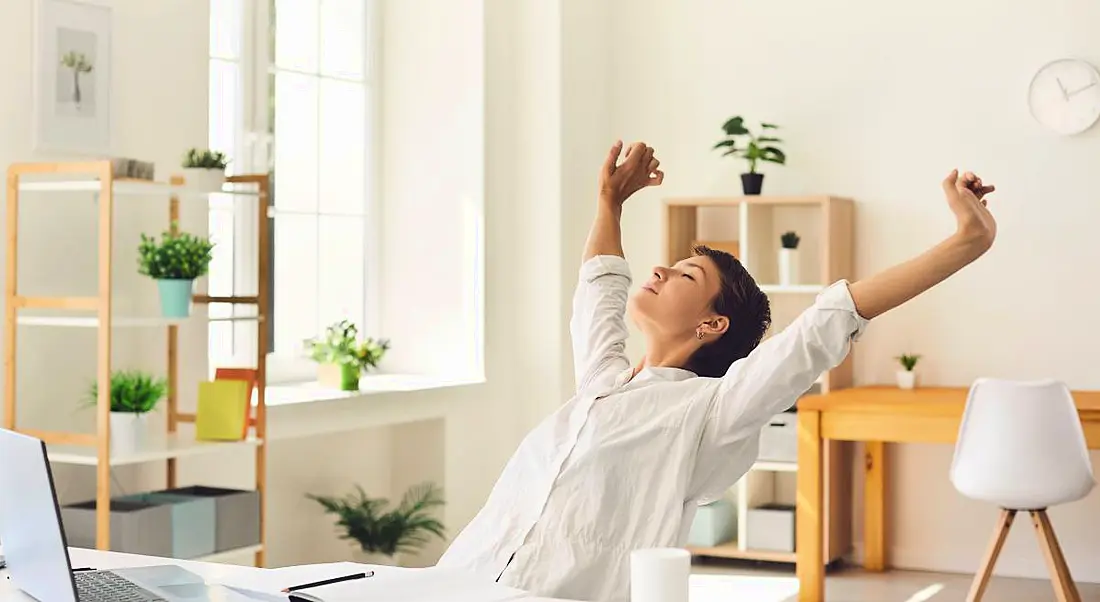 A woman working from home, stretching her arms, symbolising taking a break in a hybrid workplace.