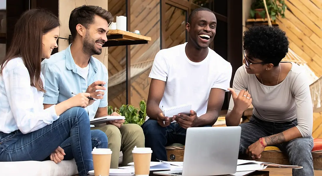 A group of young workers sitting around a laptop with coffees laughing and joking, symbolising work friendships.