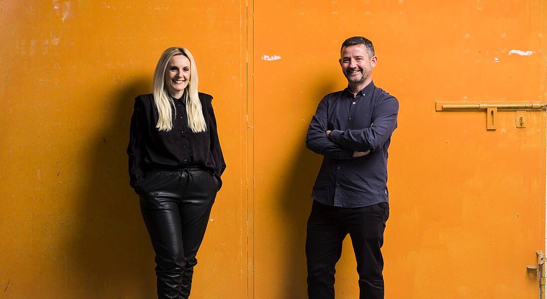 A man and a woman who work at Avcom standing in front of an orange painted wall.