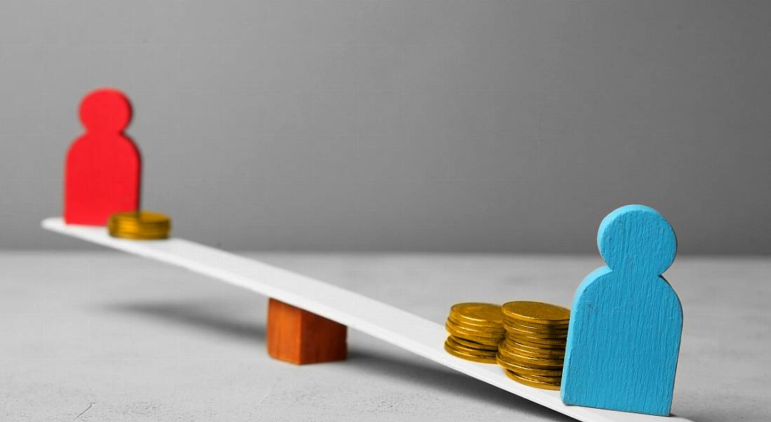 Two figures, one painted red and the other blue, on either side of a see-saw with coins on it to weigh one side down, illustrating a gender pay gap concept.