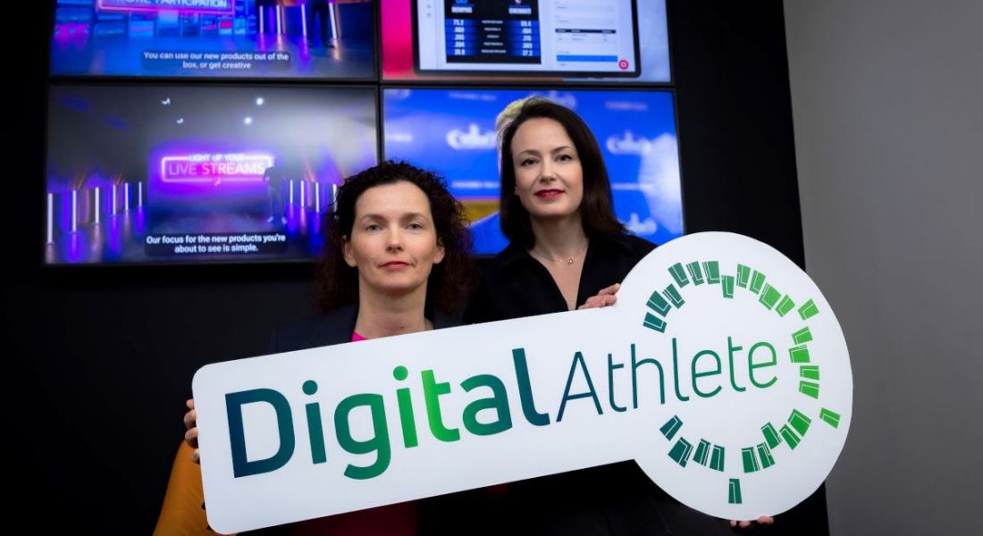 Two women holding a sign with Digital Athlete on it. They are standing in front of screens.