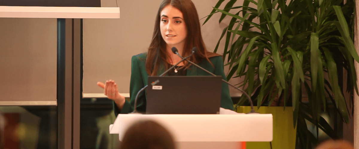 A woman wearing a green suit jacket stands behind a podium giving a speech about gender equality.