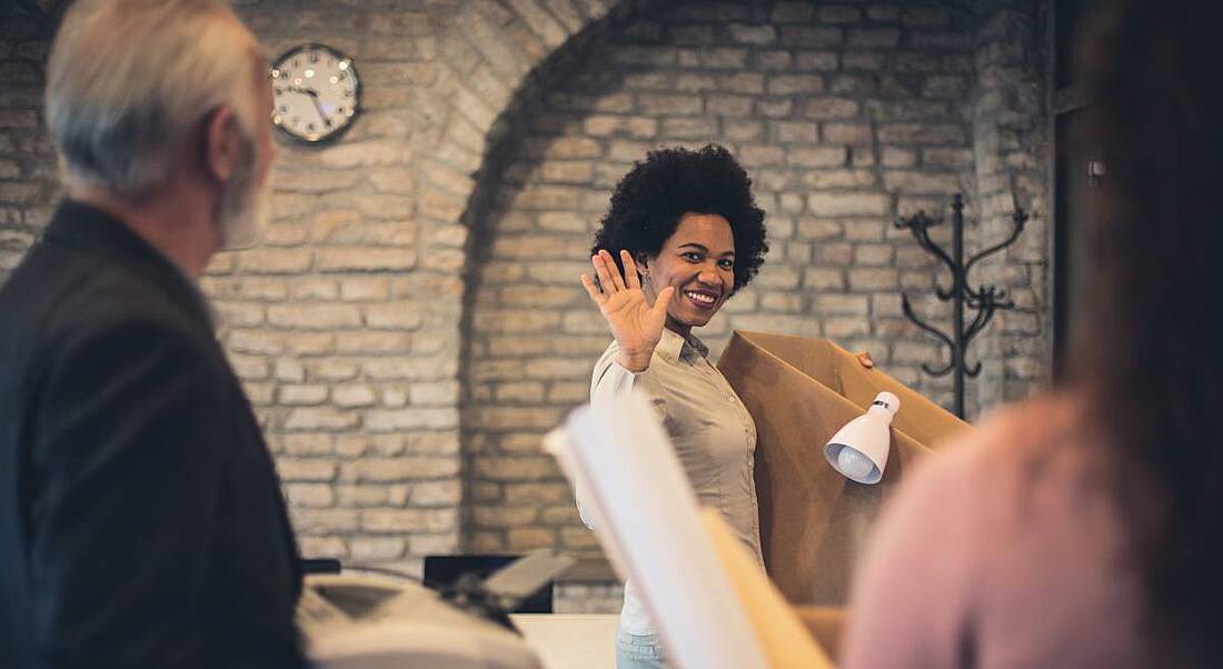 A woman holding a cardboard box, waving goodbye to her colleagues, symbolising when employees resign.