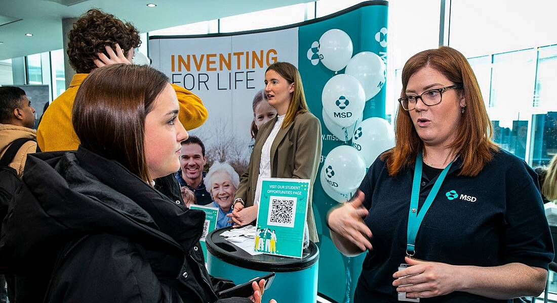 A woman wearing an MSD polo shirt speaking to a younger woman at a large career fair stand with balloons in the background.