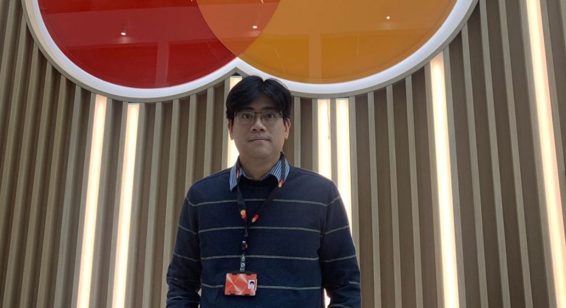 A man stands in front of a wall with the Mastercard logo on it. He is Ngoc Minh Tran, a lead data scientist at Mastercard.