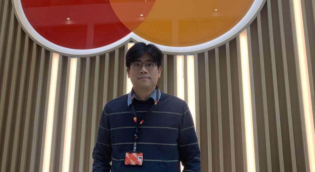 A man stands in front of a wall with the Mastercard logo on it. He is Ngoc Minh Tran, a lead data scientist at Mastercard.