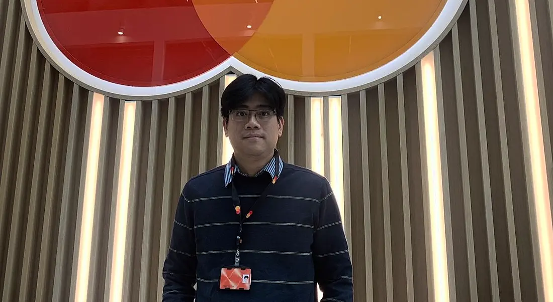 A man stands in front of a wall with the Mastercard logo on it. He is Ngoc Minh Tran, a lead data scientist at Mastercard.