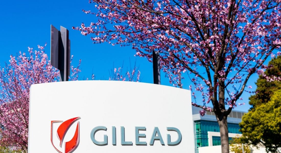 A large sign with Gilead on it for Gilead Sciences surrounded by blue sky and pink cherry blossom trees.