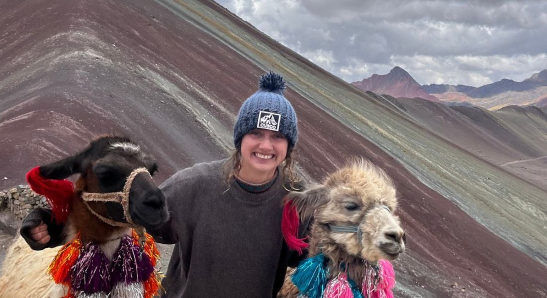 Maths teacher Alex Kelly with her arms around two decoratively dressed alpacas in front of a mountain.