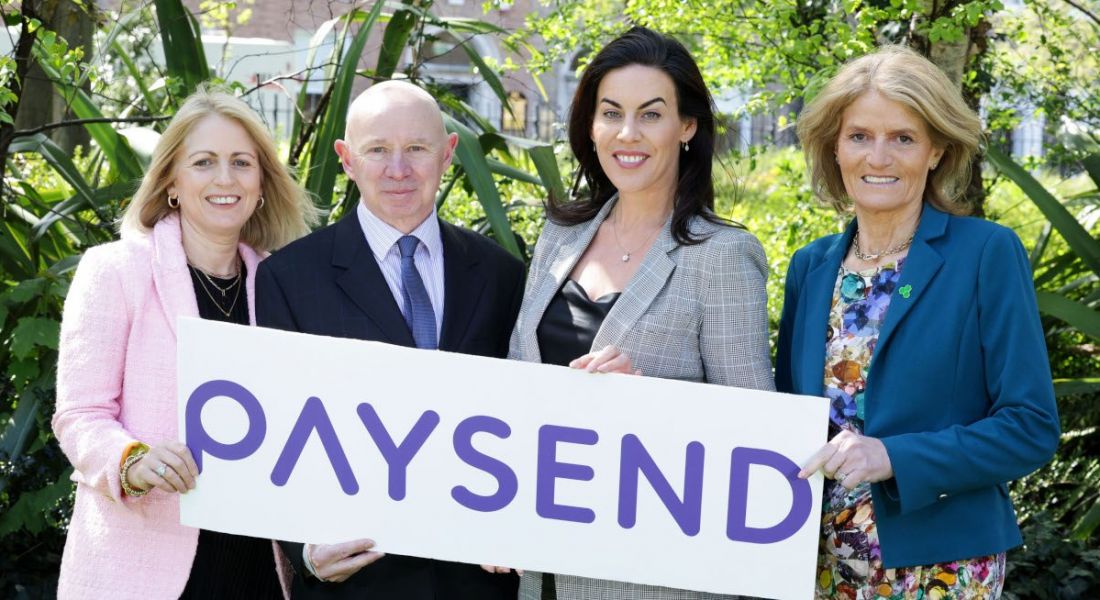 Three women and one man stand outside in business attire holding a white sign with the word 'Paysend' on it.