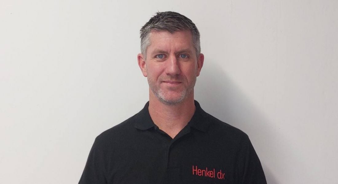 A man looks at the camera. He has short greying hair. He wears a black t-shirt with Henkel dx written on the right upper corner.