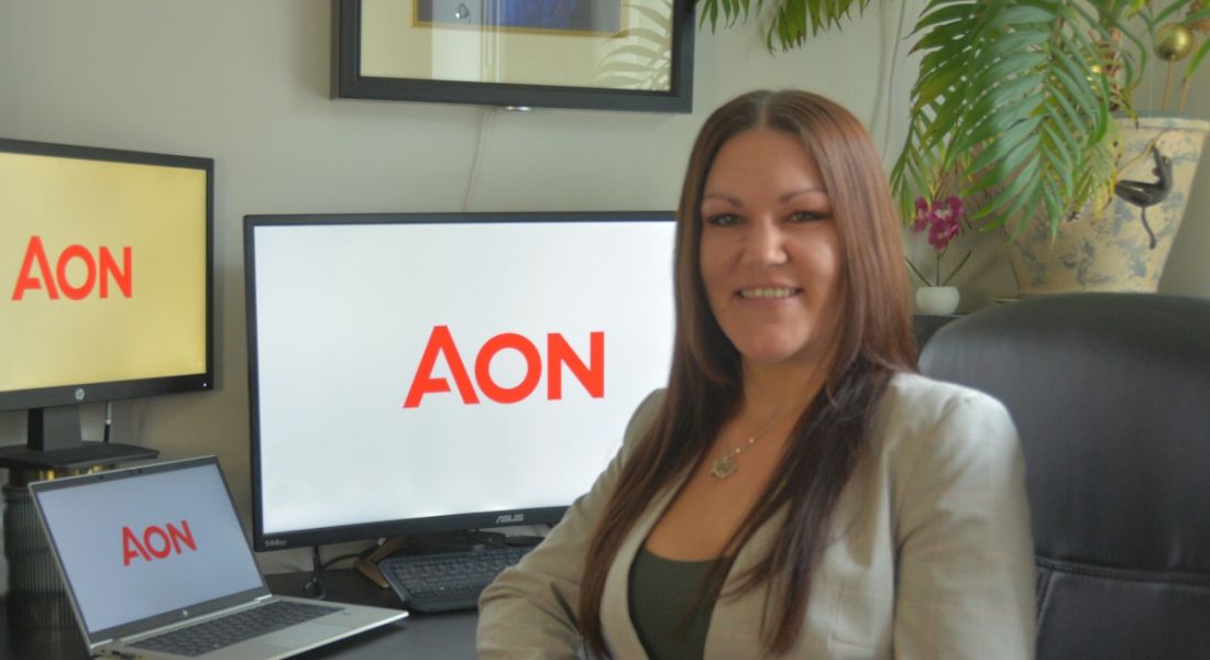 A woman sits at a desk with a laptop and monitor behind her displaying the Aon logo. She is Jenni Parry, an associate director of cyber risk at Aon.