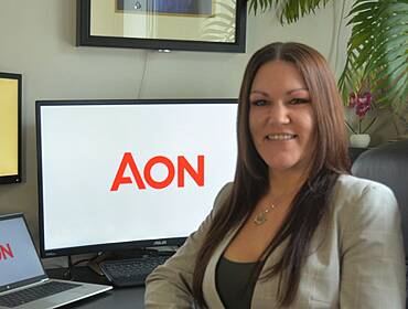 A woman sits at a desk with a laptop and monitor behind her displaying the Aon logo. She is Jenni Parry, an associate director of cyber risk at Aon.