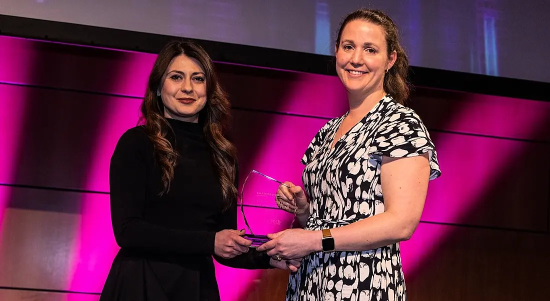 Two women stand on a large stage with purple lighting. One is presenting the other with a glass cybersecurity award.