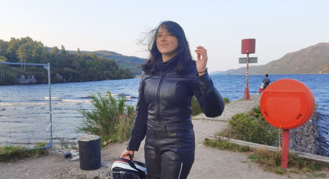 A woman wearing motorbike gear and holding a helmet against a backdrop of a body of water.