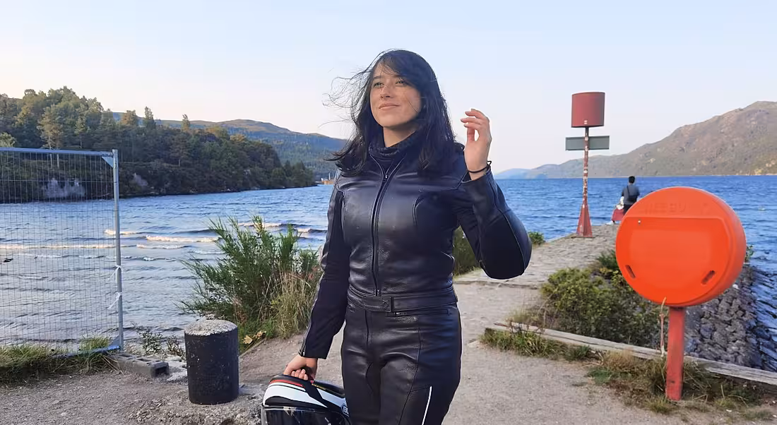 A woman wearing motorbike gear and holding a helmet against a backdrop of a body of water.