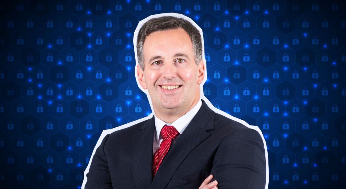 A man wearing a suit with a red tie stands smiling with his arms folded, with a blue digital background behind him.