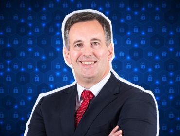 A man wearing a suit with a red tie stands smiling with his arms folded, with a blue digital background behind him.